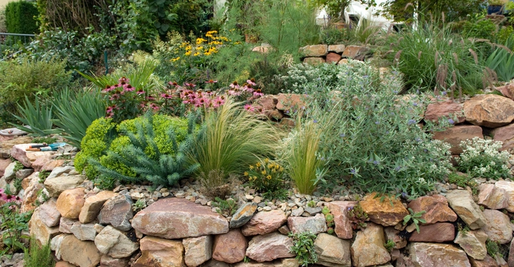 Steingarten mit bunten Blumen und Gräsern ist terrassenförmig angelegt. Verschiedene Pflanzenarten wachsen zwischen Feldsteinen. Im Hintergrund sind einige Sträucher und ein Gebäude zu sehen. Werkzeuge liegen auf den Steinen.
