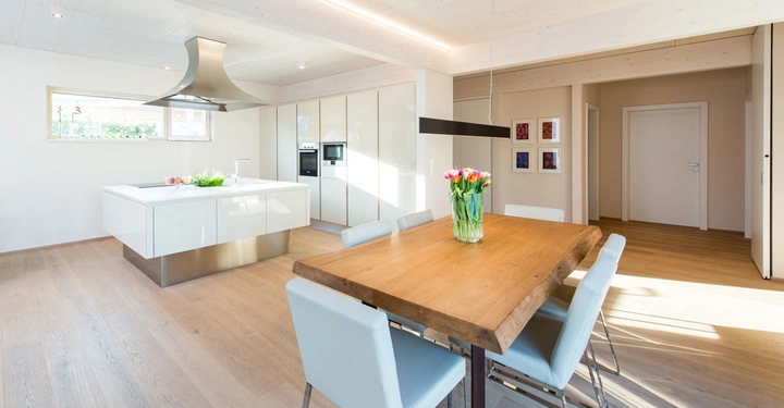 Holztisch mit einem Blumenstrauß in einem modernen Esszimmer neben einer weißen, minimalistischen Küche mit Insel und großen Fenstern, eingebettet in einem offenen, lichtdurchfluteten Raum.