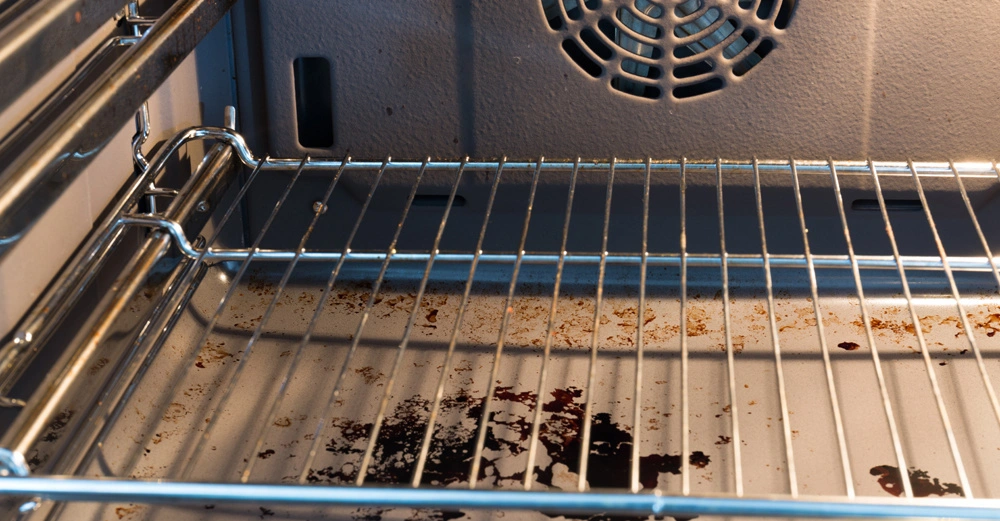 Ein Ofenrost (Objekt) mit Essensresten und Schmutz (Aktion) befindet sich in einem sauberen Ofeninnenraum (Kontext).