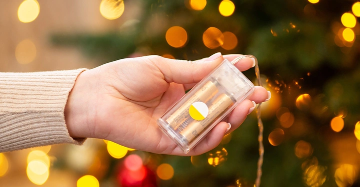 Hand hält Batteriefach einer batteriebetriebenen LED-Lichterkette vor einem Weihnachtsbaum, kabellose Weihnachtsbeleuchtung.