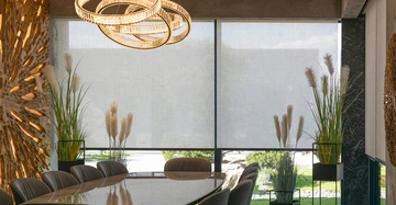 elegantes Wohnzimmer mit goldener, kunstvoller Deckenleuchte, viele Pflanzen am Fenster, Tisch und Sessel im Vordergrund angeschnitten, grau-transparente Rollos heruntergezogen