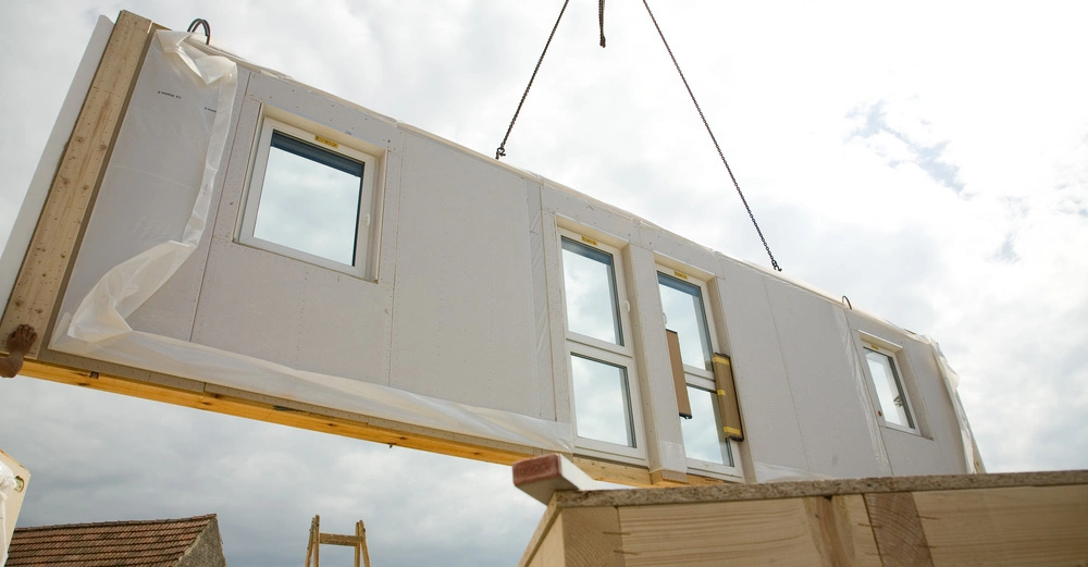 Ein großes vorgefertigtes Wandpanel mit Fenstern wird von einem Kran gehoben Baustellenumgebung mit Leiter und teilweise gebauten Strukturen unter bewölktem Himmel.
