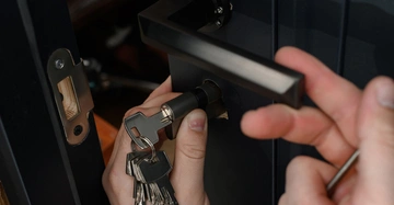 Nahaufnahme von Händen. Ein Monteur montiert ein Türschloss an einer schwarzen Eingangstür.