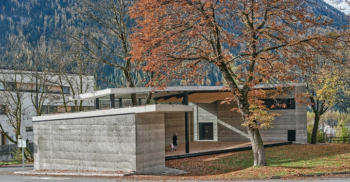 Betongebäude das am Rande eines Hofes in einer bergigen und bewaldeten Umgebung steht Zwei Personen spazieren unter einem Baum mit herbstlichen Blättern und daneben sind Gebäude sichtbar