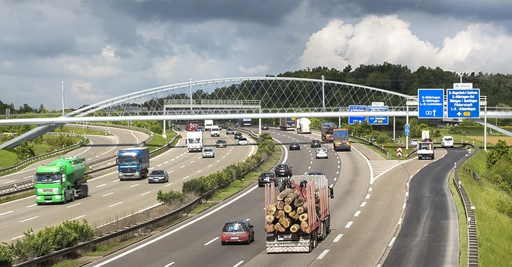 Galerie-Bild 4: Mehrspurige Autobahn mit mehreren Fahrzeugen darunter ein Lastwagen beladen mit Baumstämmen eine Fußgängerbrücke überquert die Autobahn Straßenschilder auf Deutsch leiten die Verkehrsteilnehmer dichte Wolken am Himmel.