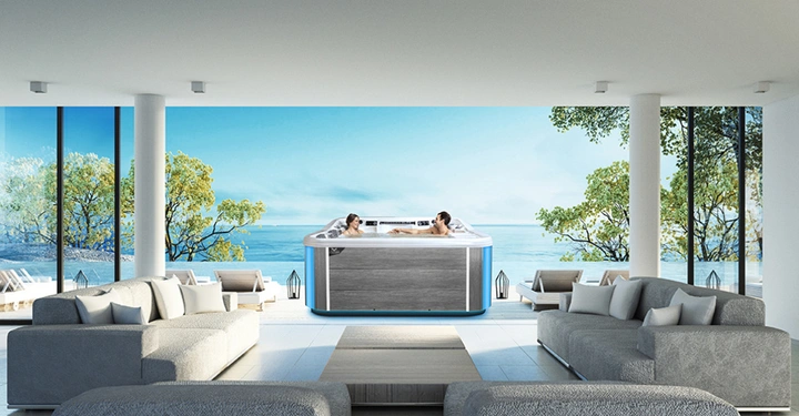 Zwei Menschen entspannen sich in einem großen Whirlpool in einem eleganten Wohnbereich mit grauen Sofas am Meer mit großen Fenstern, die Blick auf Bäume und das Wasser bieten.