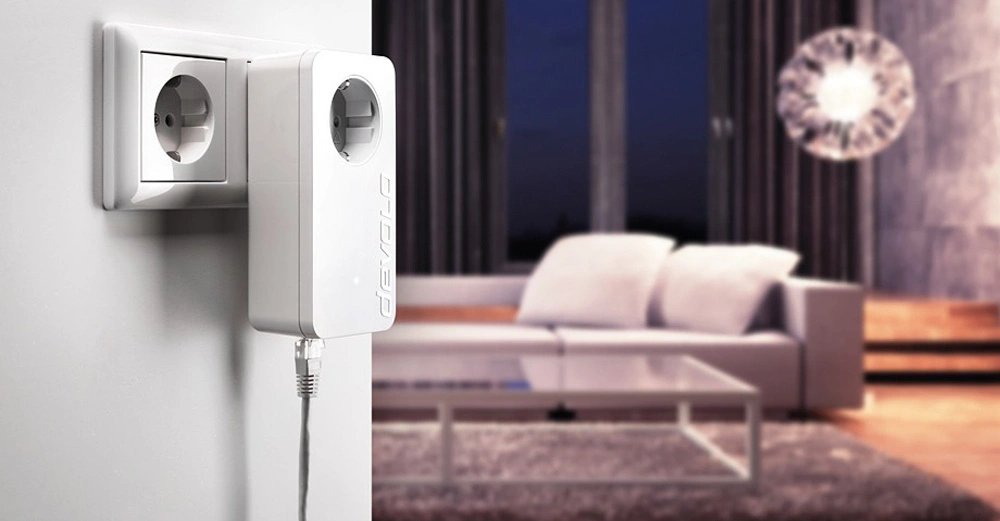 Ein weißer Steckdosenadapter mit der Aufschrift devolo ist in eine Wandsteckdose eingesteckt und zeigt nach unten ein angeschlossenes Kabel in einem modernen Wohnzimmer mit Couch und Glastisch.