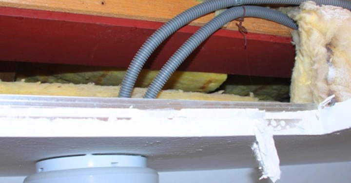 Galerie-Bild 21: Graue Kabel verlaufen durch eine offene Decke mit sichtbaren Dämmstoffen und Holzbalken. Eine montierte Leuchte befindet sich an der weißen Decke mit abblätternder Farbe.