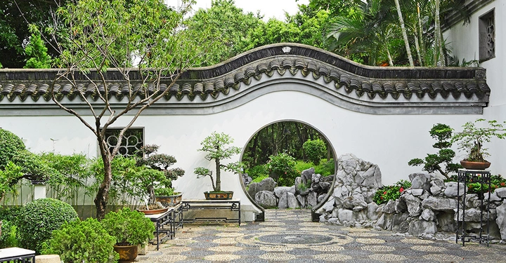 japanischer Garten mit gewundener weißer Betonmauer und gedecktem Dach. Runder Durchgang, im Vordergrund gepflasterter Weg, typisch japanische Sträucher