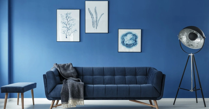 großes indigoblaues Sofa vor blauer Wand mit weißen Bildern. Rechts im Bild eine große silberne Stehlampe im Retrostil, links ein blauer tiefer Lehnsessel.