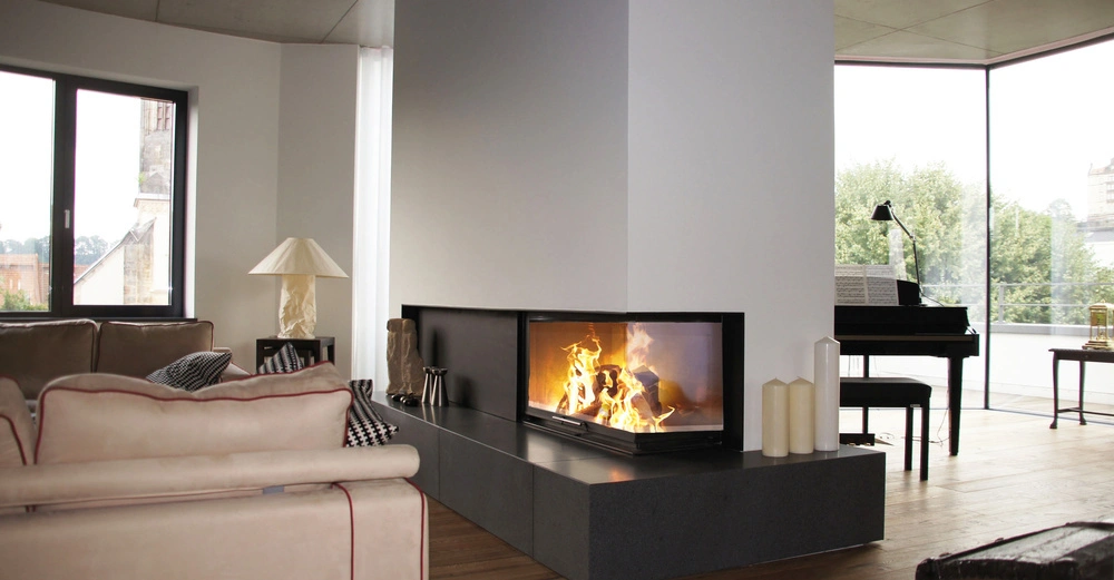 Ein Kamin feuert in einem modernen Wohnzimmer neben großen Fenstern die eine grüne Außensicht bieten Ein beiges Sofa und ein Klavier befinden sich im Raum