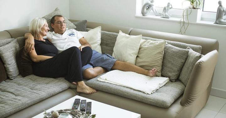 Ein Paar sitzt eng umschlungen auf einem großen grauen Sofa zahlreicher Kissen vor einem niedrigen weißen Tisch mit Fernbedienungen und Dekoration in einem hellen Wohnzimmer mit Pflanzen und Skulpturen.