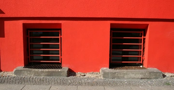 Zwei rechteckige Kellerfenster mit roten Gitterstäben befinden sich nebeneinander in einer kräftig roten Wand auf Straßenniveau mit grauem Pflaster.