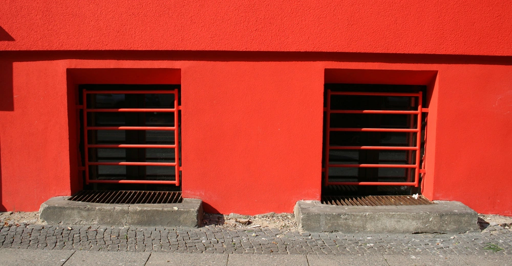 Zwei rechteckige Kellerfenster mit roten Gitterstäben befinden sich nebeneinander in einer kräftig roten Wand auf Straßenniveau mit grauem Pflaster.