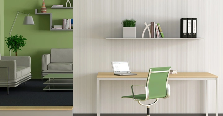 Schreibtisch mit grünem Stuhl vor beiger Wand, im Hintergrund links Eingang in grün gehaltenes Wohnzimmer, mit hellgrüner Couch und Regal mit Grünpflanzen an der Wand