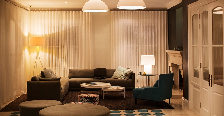 warm beleuchtetes Wohnzimmer mit Spots, Hängeleuchten und Tischlampe. Vorhänge sind zugezogen, im Vordergrund ein großes schwarzes Sofa