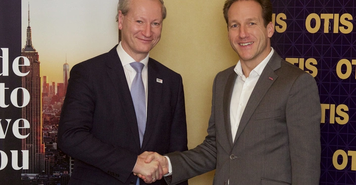 Zwei Männer schütteln sich die Hände in einem formellen Kontext. Links ist eine Stadtsilhouette und Text sichtbar Made to move you. Rechts im Hintergrund das Logo OTIS.