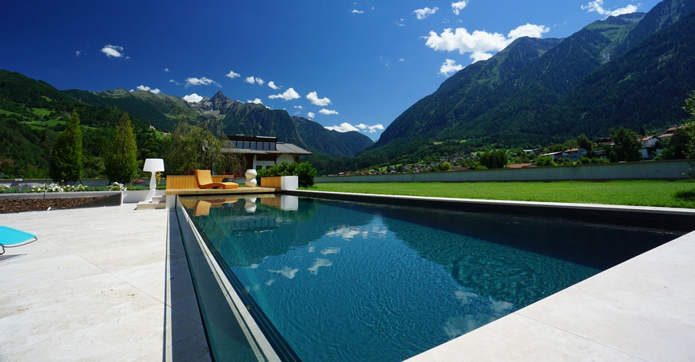 Ein rechteckiger Swimmingpool reflektiert den blauen Himmel die Sonnenterrasse hat bunte Liegen und eine Lampe die Umgebung besteht aus grünen Wiesen und hohen Bergen im Hintergrund