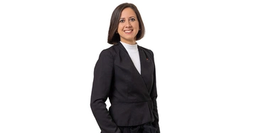 Frau lächelt und trägt einen schwarzen Blazer und einen weißen Rollkragenpullover steht vor einem weißen Hintergrund