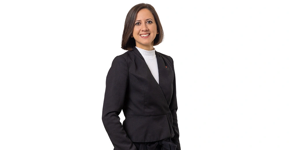 Frau lächelt und trägt einen schwarzen Blazer und einen weißen Rollkragenpullover steht vor einem weißen Hintergrund