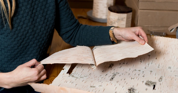 Eine Person blättert in handgefertigtem Papier an einem Holztisch Umgebung mit weiteren Papierrollen und Kartons im Hintergrund.