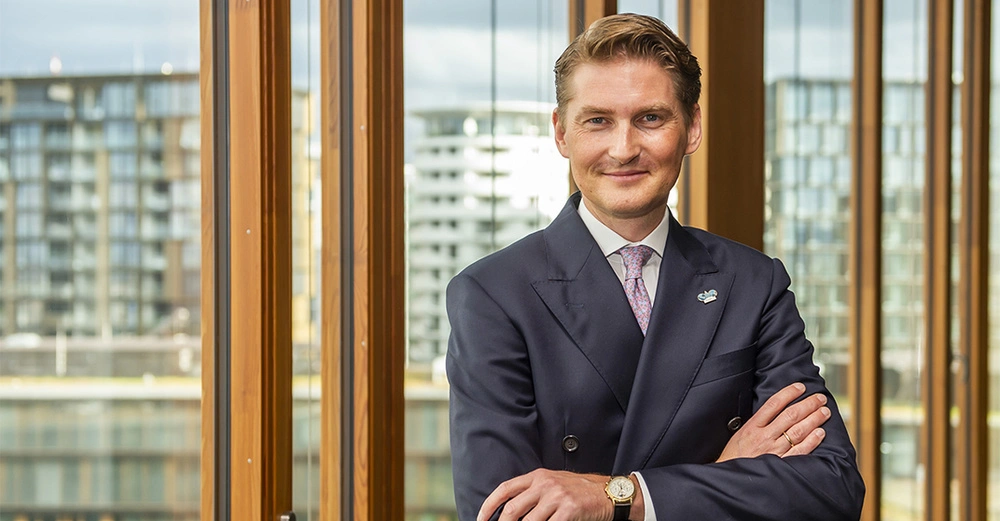 Ein Mann trägt einen Anzug und steht mit verschränkten Armen in einem modernen Gebäude vor großen Fenstern, mit einer städtischen Landschaft und hohen Gebäuden im Hintergrund.