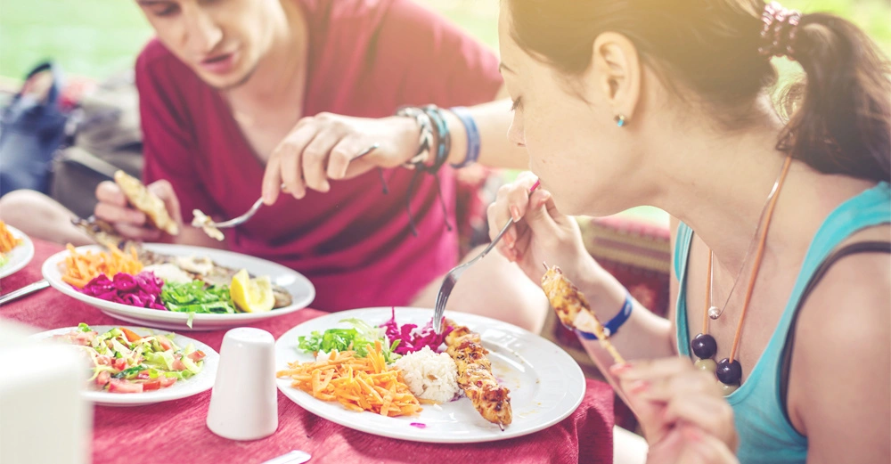 Zwei Personen essen bunte Salate und gegrilltes Gemüse an einem Tisch im Freien an einem warmen sonnigen Tag umgeben von viel Grün.