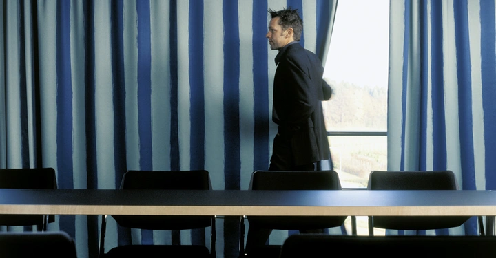 Galerie-Bild 15: Eine Person im Anzug zieht blaue Vorhänge in einem Konferenzraum auf wo schwarze Stühle hinter einem langen Tisch stehen und Licht durch das Fenster fällt.