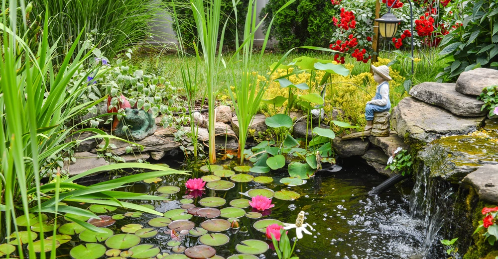 Statue sitzt am Teichrand neben Wasserfall und Seerosen im blühenden Garten mit hohen Gräsern und roten Blumen im Hintergrund