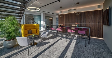 Gelber Sessel und graue Stühle stehen um einen Tisch in einem modernen Büro mit Treppe im Hintergrund gepunktetem Teppichmuster grafischer Holztrennwand und rosa Barhockern an einem Hochstehtisch.