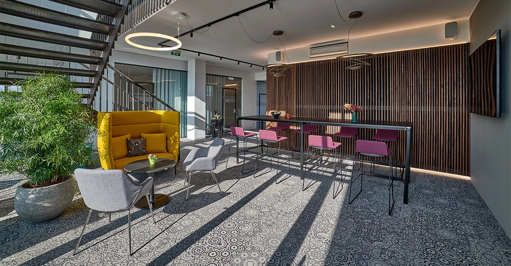 Gelber Sessel und graue Stühle stehen um einen Tisch in einem modernen Büro mit Treppe im Hintergrund gepunktetem Teppichmuster grafischer Holztrennwand und rosa Barhockern an einem Hochstehtisch.