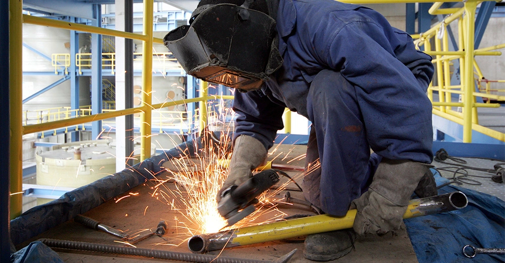 Person in schwerer Arbeitskleidung und Schweißerhelm kniet auf einem Metallgerüst und schneidet ein Rohr mit einer Kreissäge, während Funken sprühen.