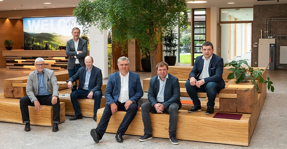 Sechs Männer in Anzügen sitzen und stehen auf Holzbänken eine große Pflanze daneben im modernen Bürogebäude mit WILLKOMMEN-Logo und Rezeption im Hintergrund