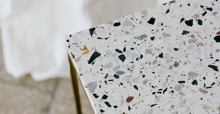 Ecke eines Tisches aus Terrazzo-Material zeigt schwarze, graue und beige Marmorstücke in weißem Zement eingebettet stehend in einem hellen Raum auf gemustertem Boden.
