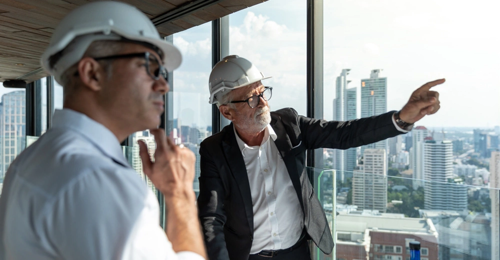 zwei Männer in weißem Hemd, einer mit schwarzem Sakko und Baustellenhelmen auf dem Kopf unterhalten sich auf der Baustelle, einer zeigt mit der Hand in die Ferne