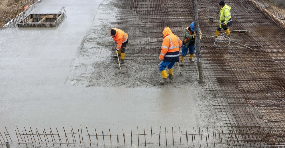 Arbeiter gießen und glätten Beton auf einer Baustelle wobei sie Sicherheitsausrüstung wie reflektierende Jacken und Gummistiefel tragen. Die Umgebung besteht aus Baustahlmatten und unvollendeten Abschnitten.