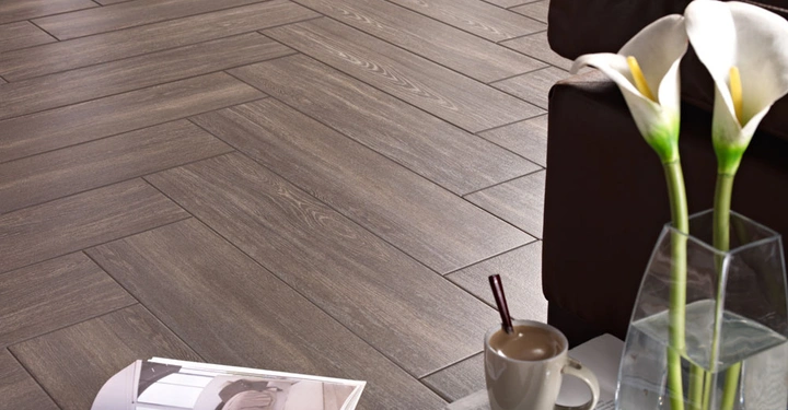 Braune Keramikfliesen bilden den Boden in einem Wohnzimmer; im Vordergrund stehen eine Vase mit weißen Lilien eine Kaffeetasse mit Löffel und ein aufgeschlagenes Buch auf einem Couchtisch.