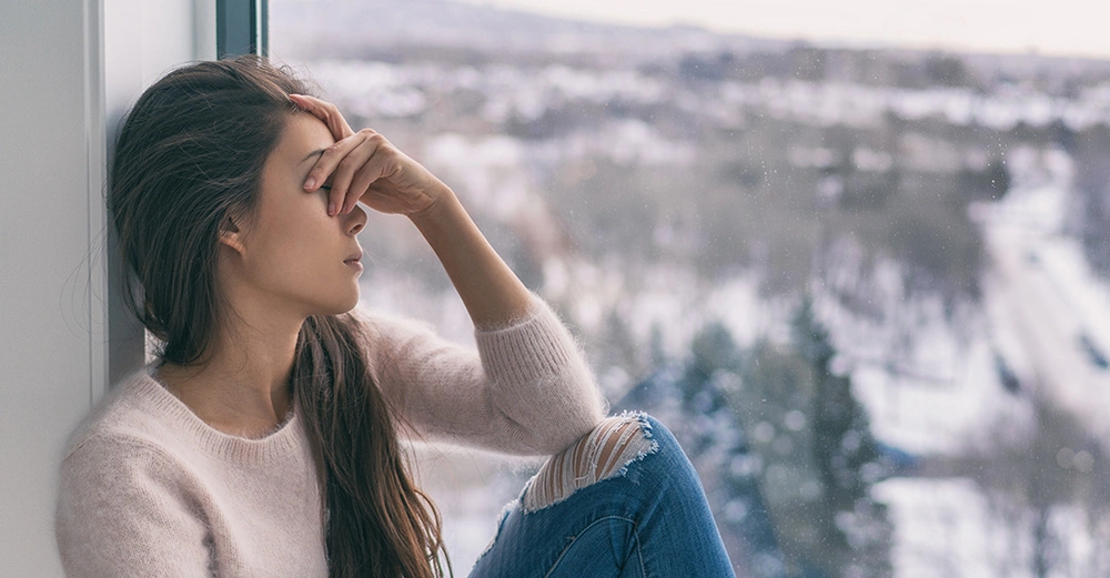 Frau mit Symptomen von Winterblues und saisonaler Depression (SAD) sitzt im Winter allein am Fenster.