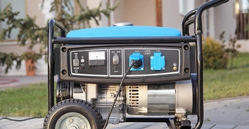 Stromgenerator steht auf einem gepflasterten Weg vor einem Haus und wird durch ein Verlängerungskabel betrieben Im Hintergrund sind Rasen und Sträucher zu sehen