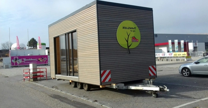 Ein kleines Holzhaus steht auf einem Anhänger auf einem Parkplatz neben einem Auto und gegenüber von einem rosa Verkaufsstand mit der Aufschrift Guschlbauer Der Sonntagskrapfen und einer weißen Energie AG Anlage. Auf dem Holzhäuschen steht HolZhaus to go
