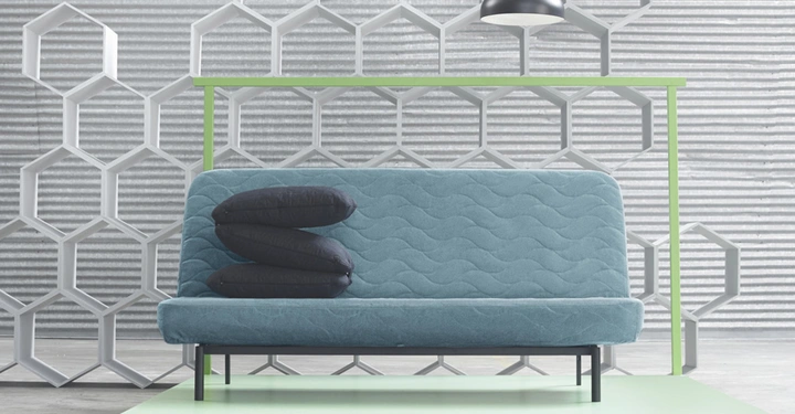Galerie-Bild 8: Ein blaues Sofa steht auf einem hellgrünen Podest, darauf liegen drei dunkelblaue Kissen übereinander. Im Hintergrund eine geometrische Musterwand und eine hängende Lampe.
