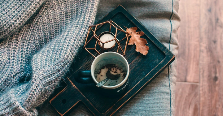 Tasse Kaffee auf Holztablett mit Blatt und Kerze neben Strickpullover auf Sofa dunkler Holzboden im Hintergrund