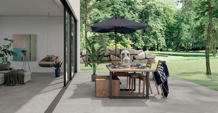 Essgruppe steht auf einer gepflasterten Terrasse mit einem Sonnenschirm darüber. Im Hintergrund eine grüne Landschaft. Nebenan befindet sich ein modernes Wohnzimmer mit Hängesessel und Pflanzen.