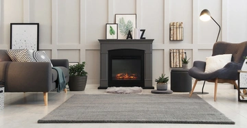 Ein grauer Kamin im modernen Wohnzimmerumfeld mit brennendem Feuer dekoriert mit Pflanzen Bildern und Buchstaben A Z umgeben von einem grauen Sessel Sofas und einem Teppich.