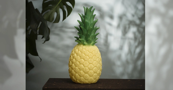 Galerie-Bild 2: Eine gelbe Ananas-Lampe steht leuchtend auf einem dunklen Holztisch im Vordergrund weiche Schatten und Monstera-Blätter im Hintergrund.