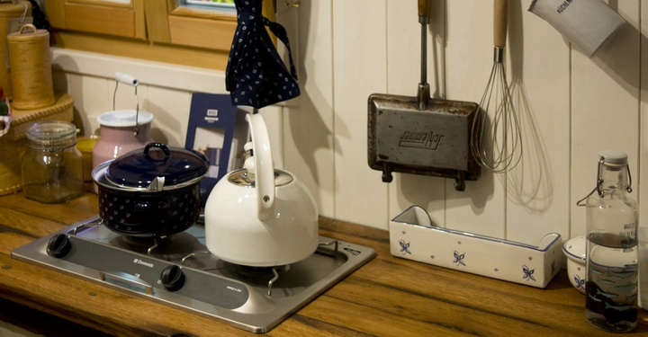 Galerie-Bild 6: Ein weißer Wasserkocher und ein blauer Topf stehen auf einem Herd in einer traditionellen Küche mit Holzmöbeln. An der Wand hängen Küchenutensilien und ein emaillierter Becher.