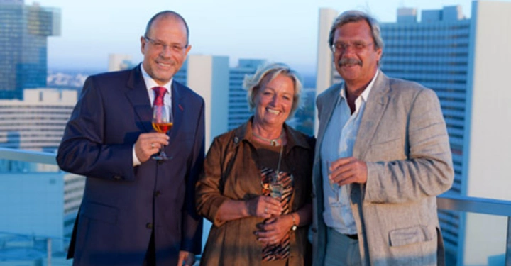 Galerie-Bild 14: Drei Menschen halten Gläser in ihren Händen und lächeln auf einem Balkon vor einer städtischen Skyline. Eine Person trägt einen Anzug, die anderen beiden sind leger gekleidet.