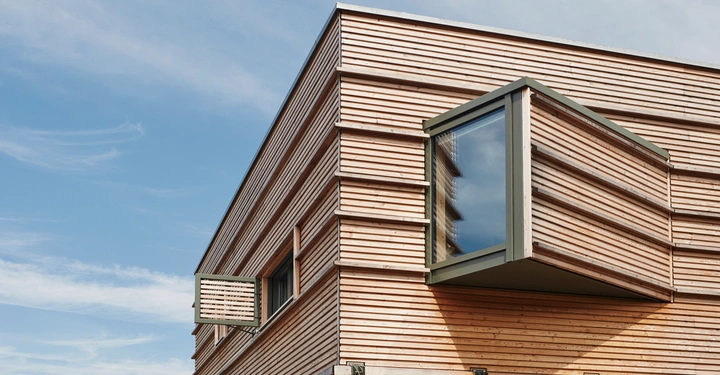 Gebäude mit hölzerner Fassade und großen, rechteckigen Fenstern ragt vor einem klaren blauen Himmel empor. Eine Fensterbox streckt sich vom Gebäude nach außen.