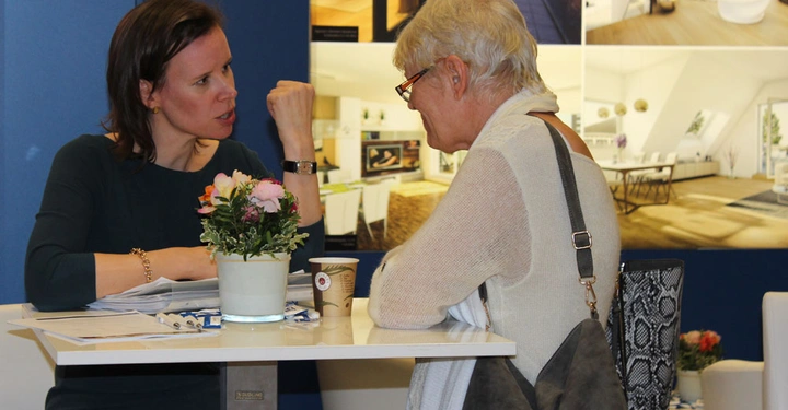 Galerie-Bild 6: Zwei Frauen sitzen an einem Tisch und unterhalten sich; eine zeigt eine erklärende Geste. Im Hintergrund sind Plakate mit Inneneinrichtungsfotos zu sehen. Auf dem Tisch steht eine Blume und eine Kaffeetasse.