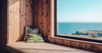 Ein Wohnzimmer mit Holzwänden und einem großen Fenster das aufs Meer blickt Ein beige Sitzsack mit zwei dekorativen Kissen in Grün und Blumenmuster steht an der Wand.